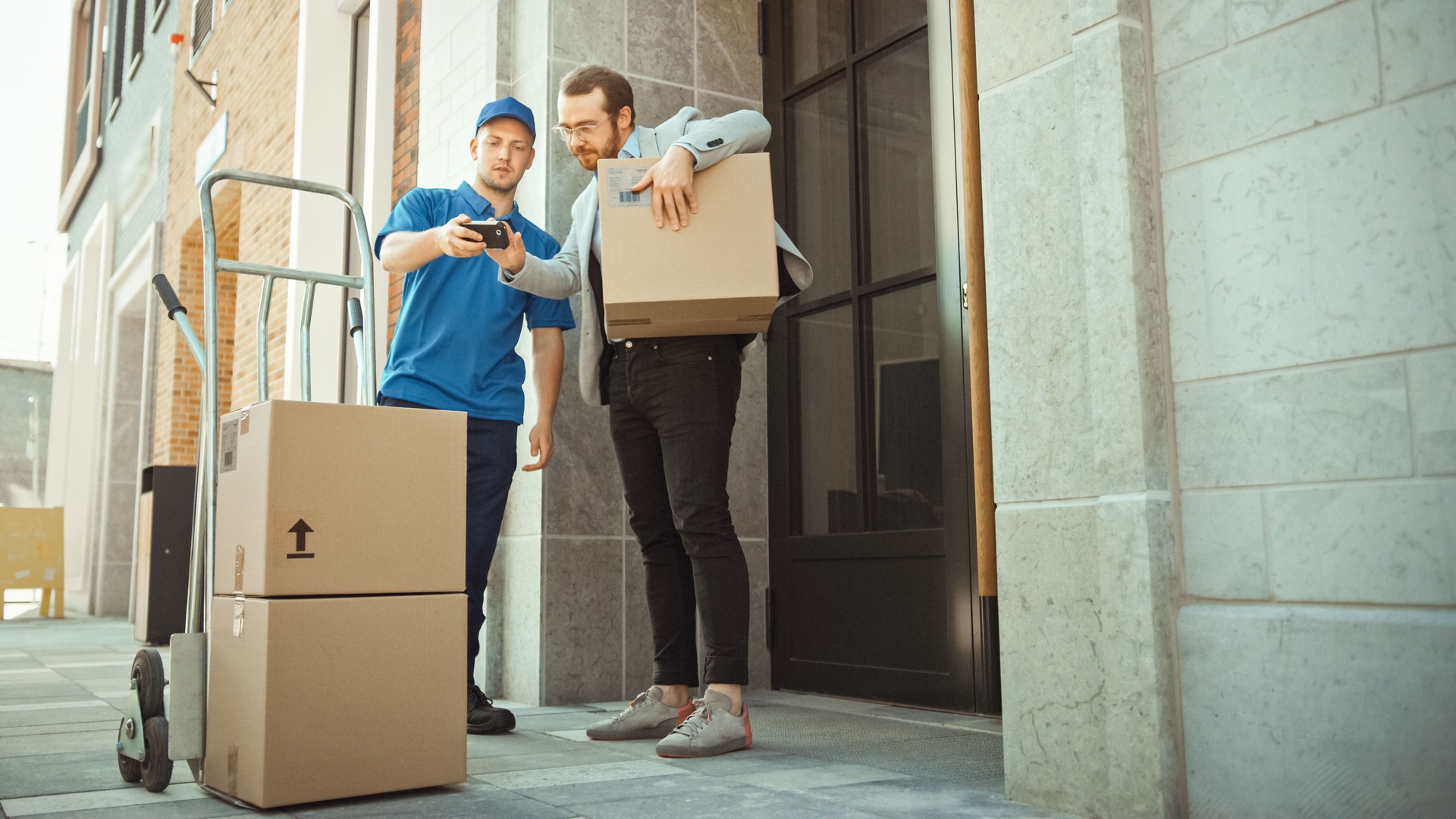 Delivery Man Pushes Hand Truck Trolley Full of Cardboard Boxes Hands Package to a Customer, Who then Signs Electronic POD Device. Courier Delivers Parcel to Man in Stylish Modern Urban Office Area